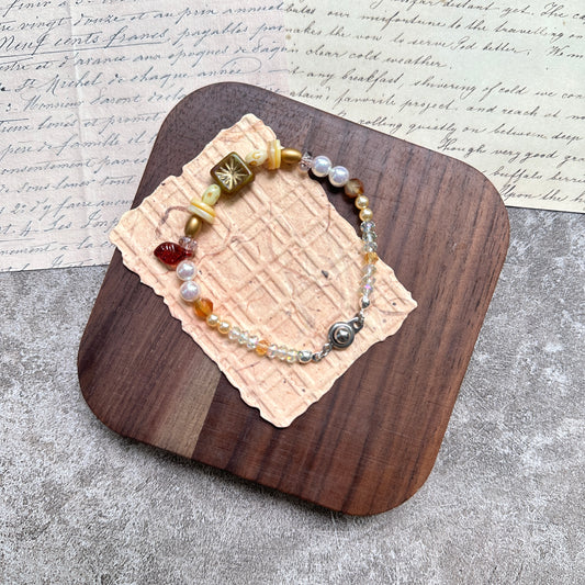 Olive Sunburst Bracelet — flat lay in a wooden tray on textured papers; honey and pearl beads with an olive square sunburst charm, red leaf accent, and magnetic clasp