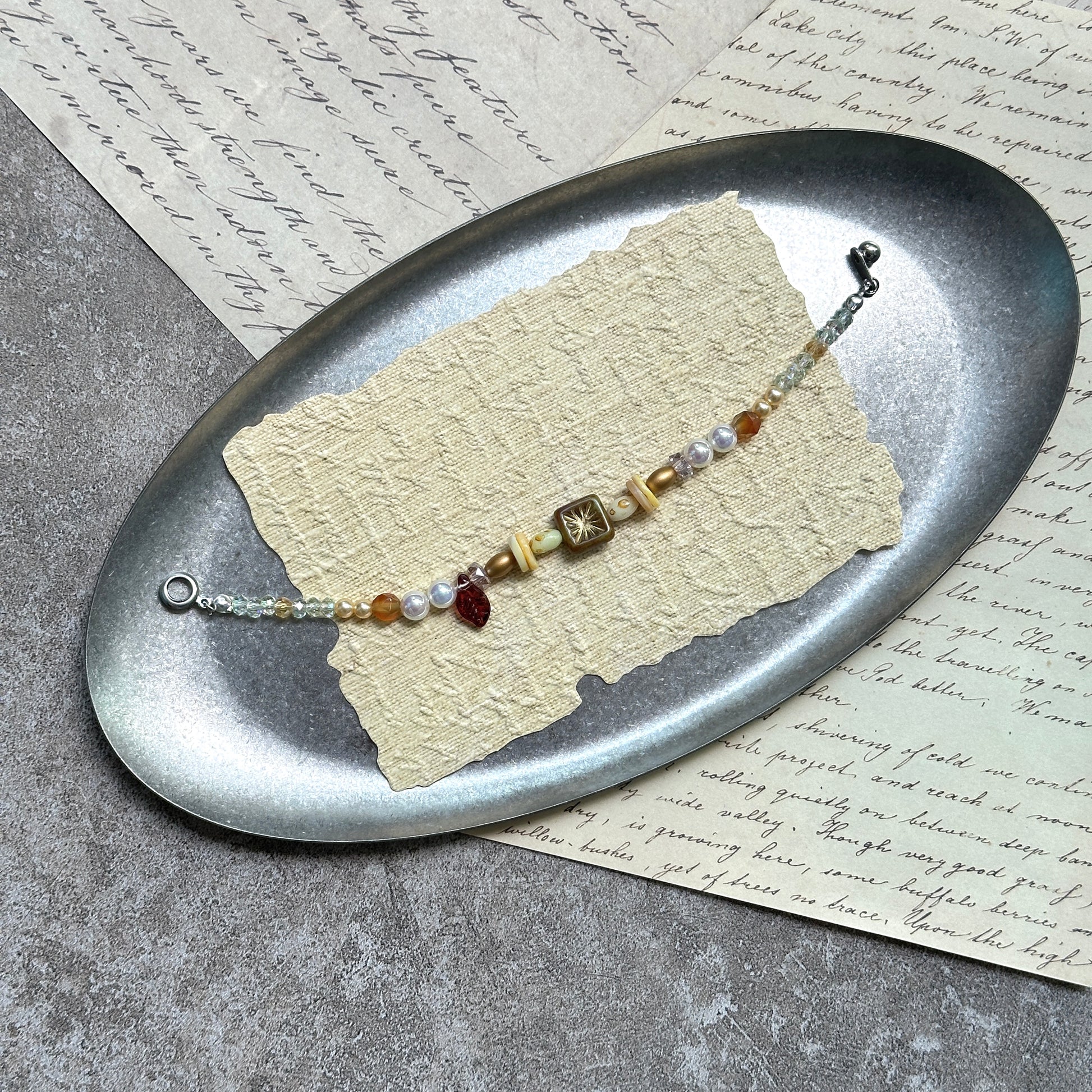Olive Sunburst Bracelet — flat lay in a wooden tray on textured papers; honey and pearl beads with an olive square sunburst charm, red leaf accent, and magnetic clasp2