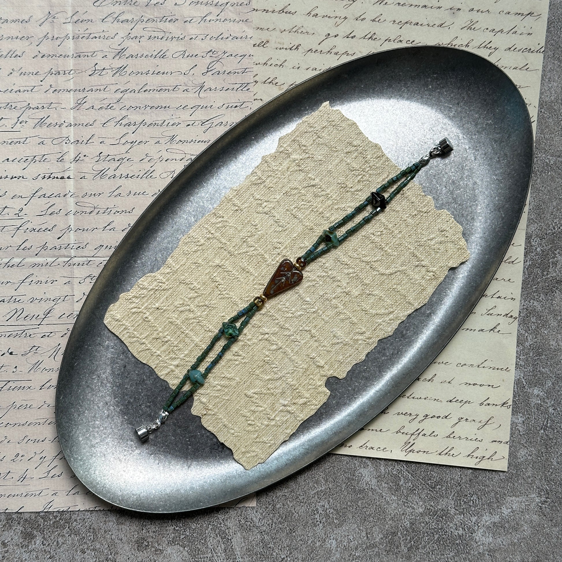 Verdant Talisman Bracelet — flat lay on textured papers; deep forest-green beads with a botanical heart talisman and magnetic clasp 2