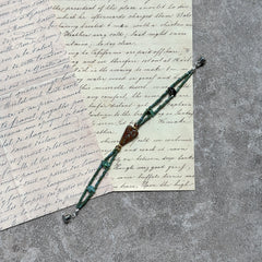 Verdant Talisman Bracelet — flat lay on textured papers; deep forest-green beads with a botanical heart talisman and magnetic clasp 3