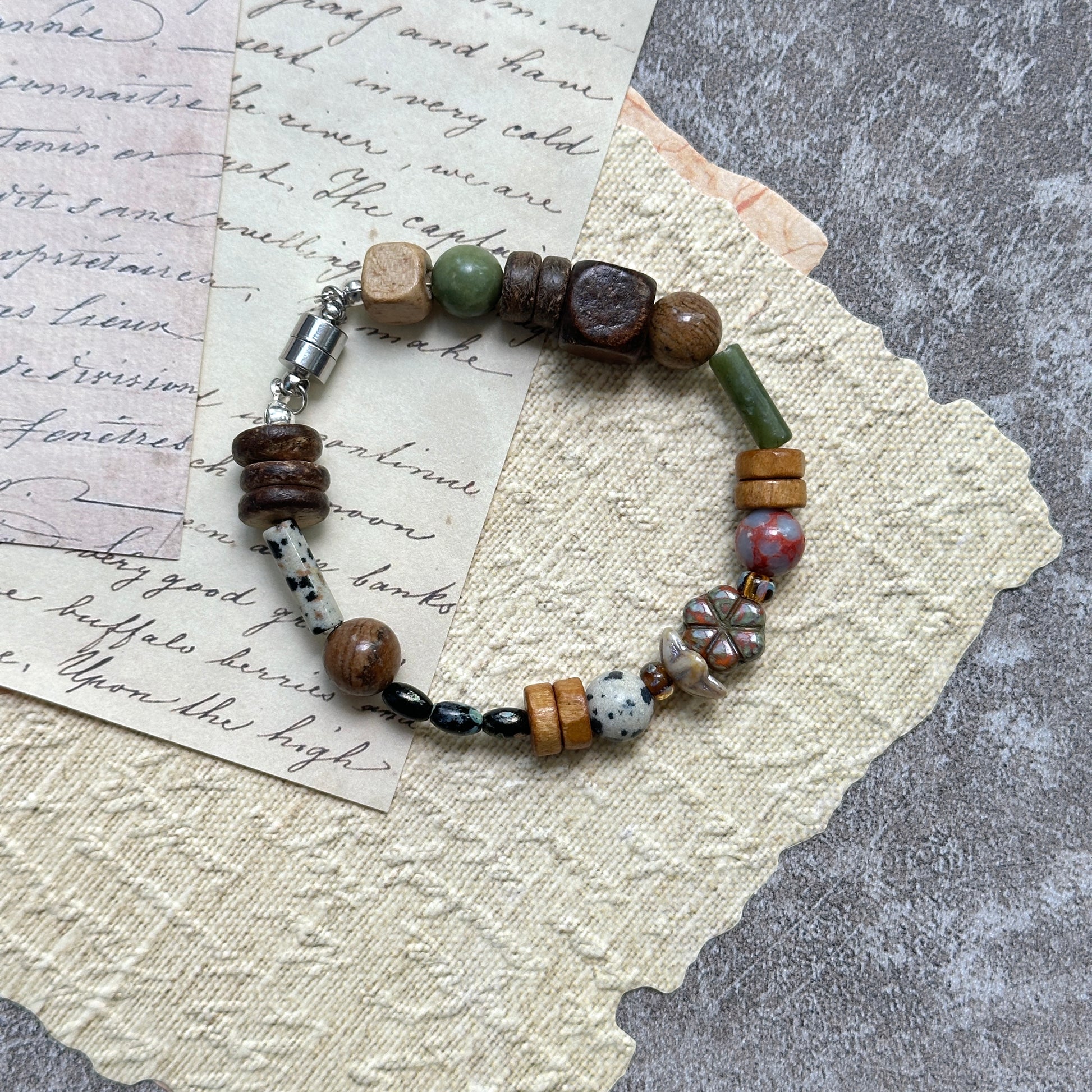Cedar & Sage Bracelet — flat lay on textured papers and wooden tray; cedar-brown and sage-green beads with speckled jasper accents and a magnetic clasp 2