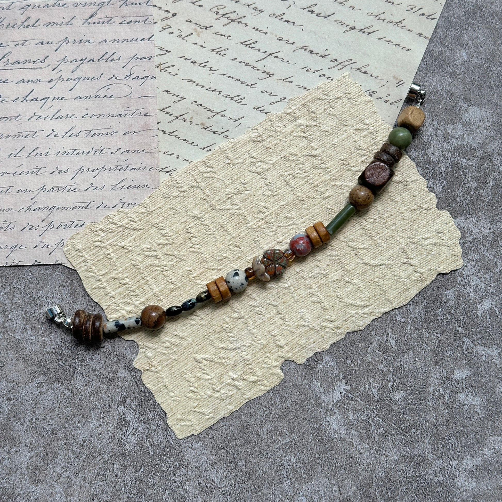 Cedar & Sage Bracelet — flat lay on textured papers and wooden tray; cedar-brown and sage-green beads with speckled jasper accents and a magnetic clasp 3