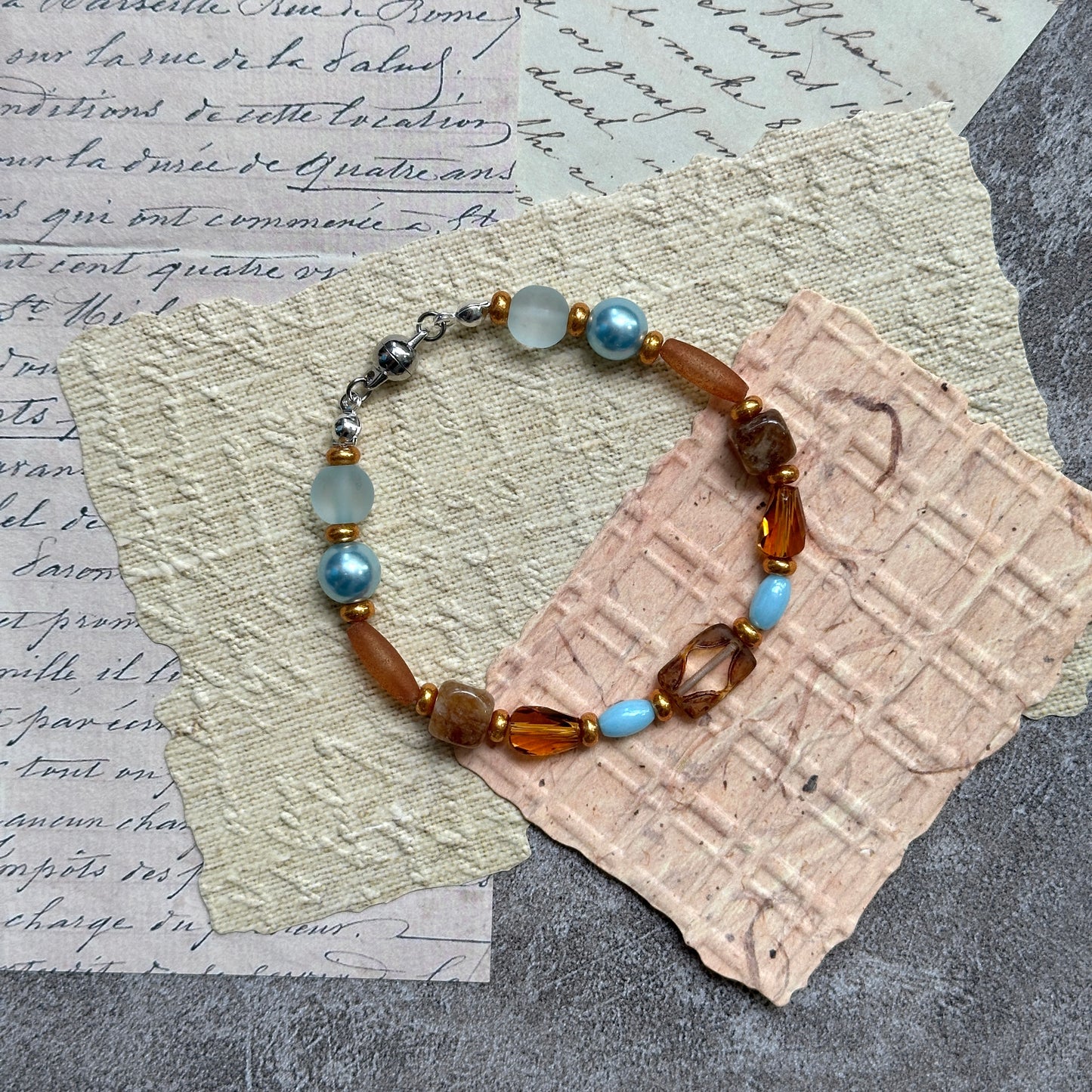 Aqua & Amber Drift Bracelet — flat lay on textured stone paper; sea-glass aqua and warm amber beads with gold accents and a nickel-free magnetic clasp 2