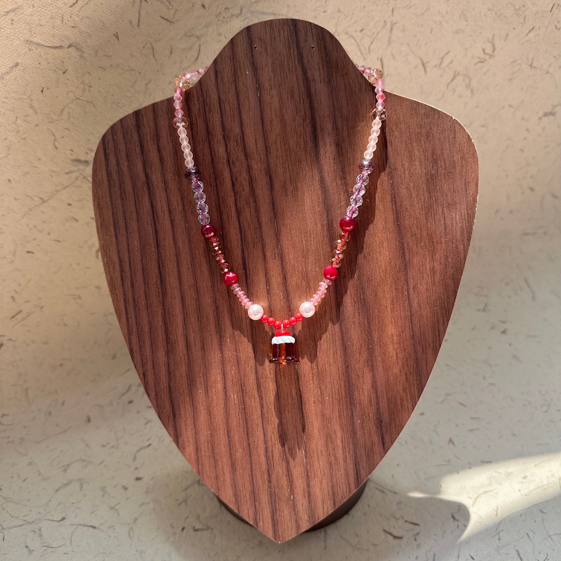 Crimson Bell Necklace on wood display bust — crimson and blush glass beads with a lampwork bell pendant