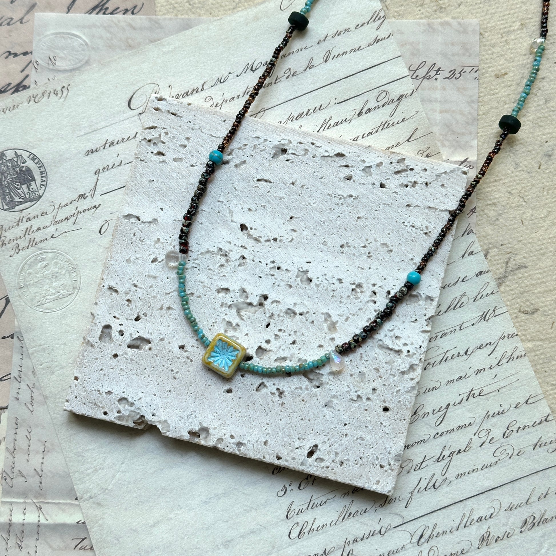 Azure Blossom Necklace — flat lay on vintage papers; double-strand bronze-toned and turquoise beads with a floral ceramic charm
