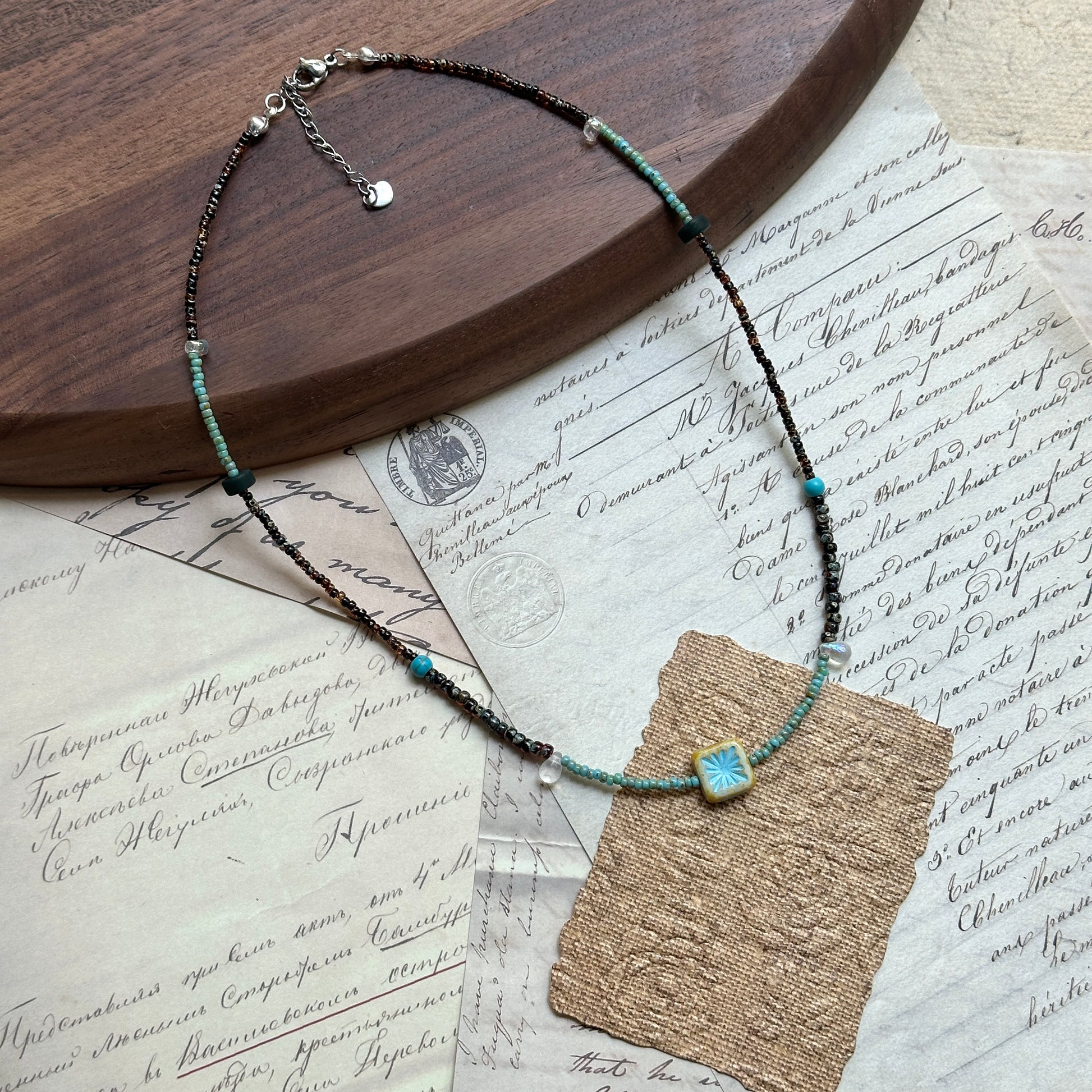 Azure Blossom Necklace — angled view beside a wooden tray; bronze-toned and turquoise beads with floral ceramic focal charm
