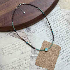 Azure Blossom Necklace — angled view beside a wooden tray; bronze-toned and turquoise beads with floral ceramic focal charm