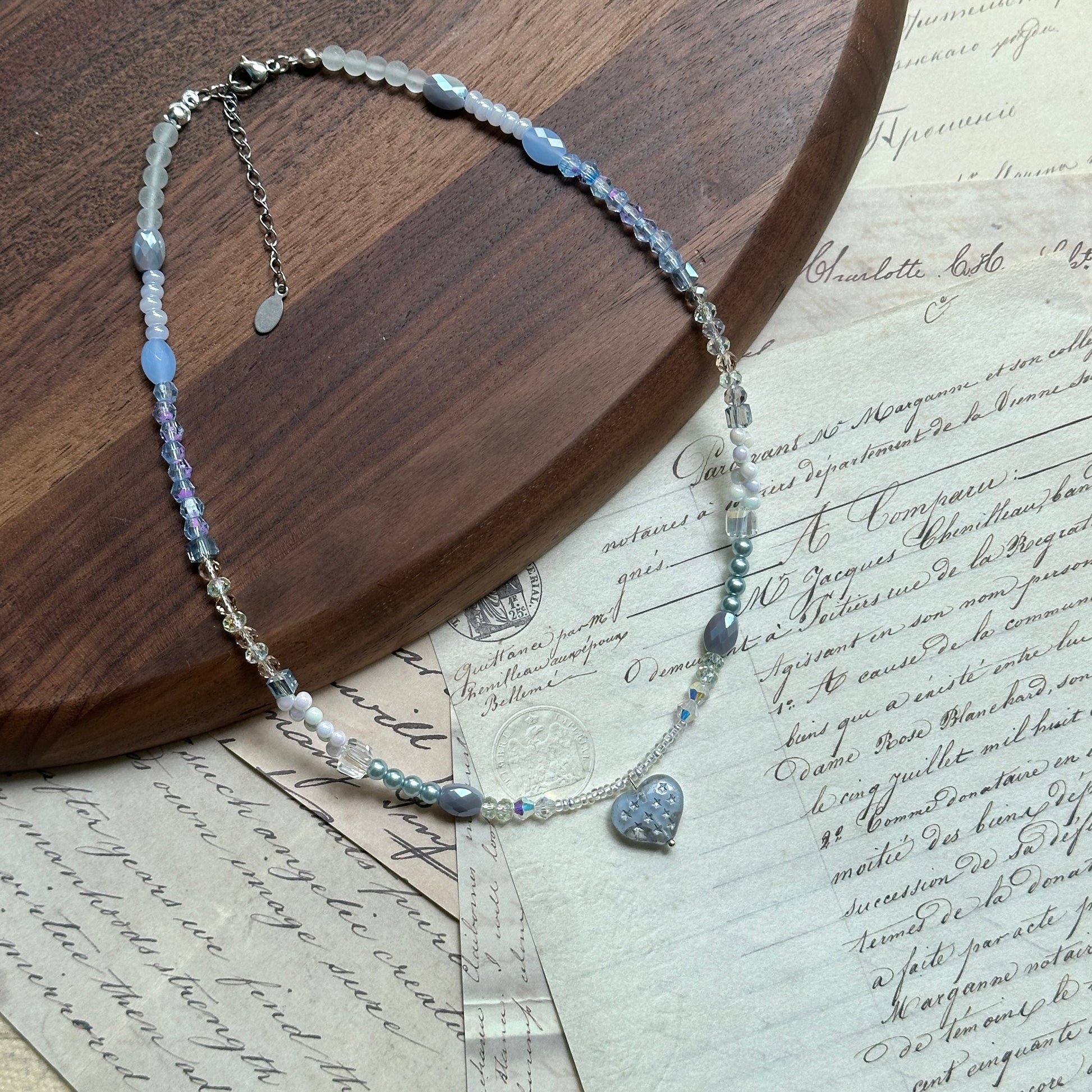 Moonlit Heart Necklace — flat lay on vintage papers beside a round wooden tray; pearl-white and icy-blue beads with a heart-shaped pendant