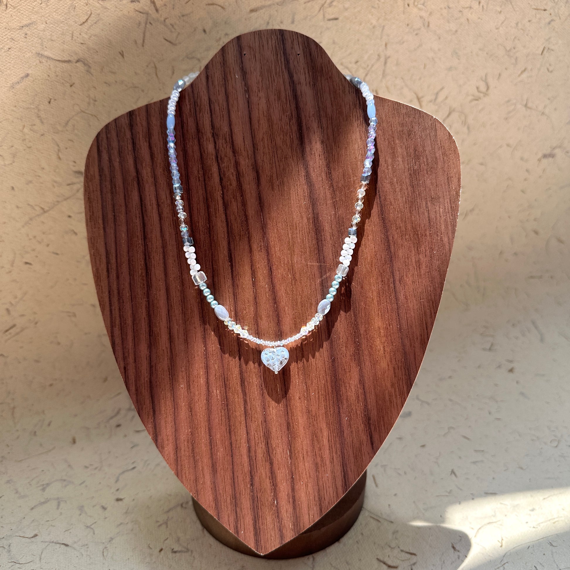 Moonlit Heart Necklace on wood display bust — pearl-white and icy-blue beads with a heart-shaped pendant