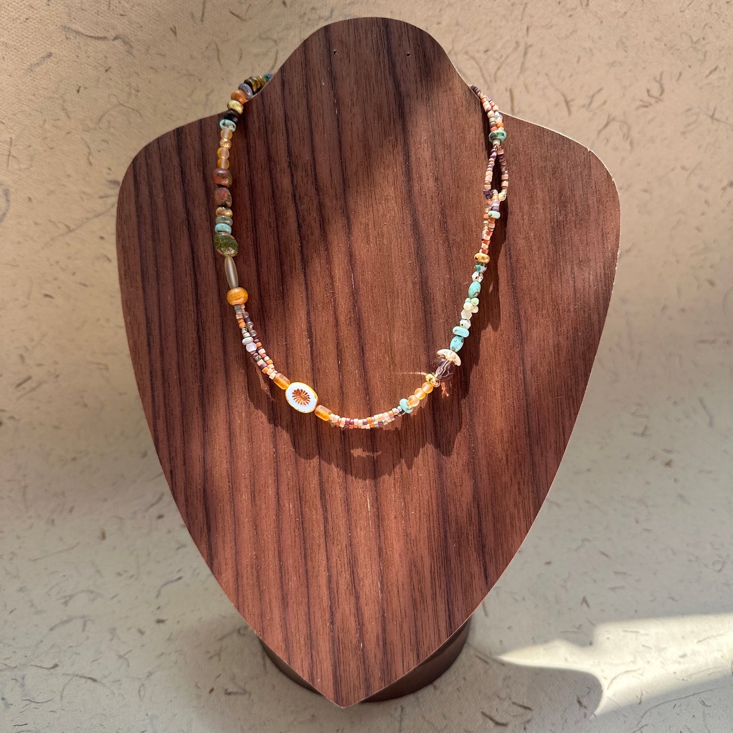 Sunlit Meadow Necklace on wood display bust — warm amber and earthy-green beads with a sunflower-inspired charm