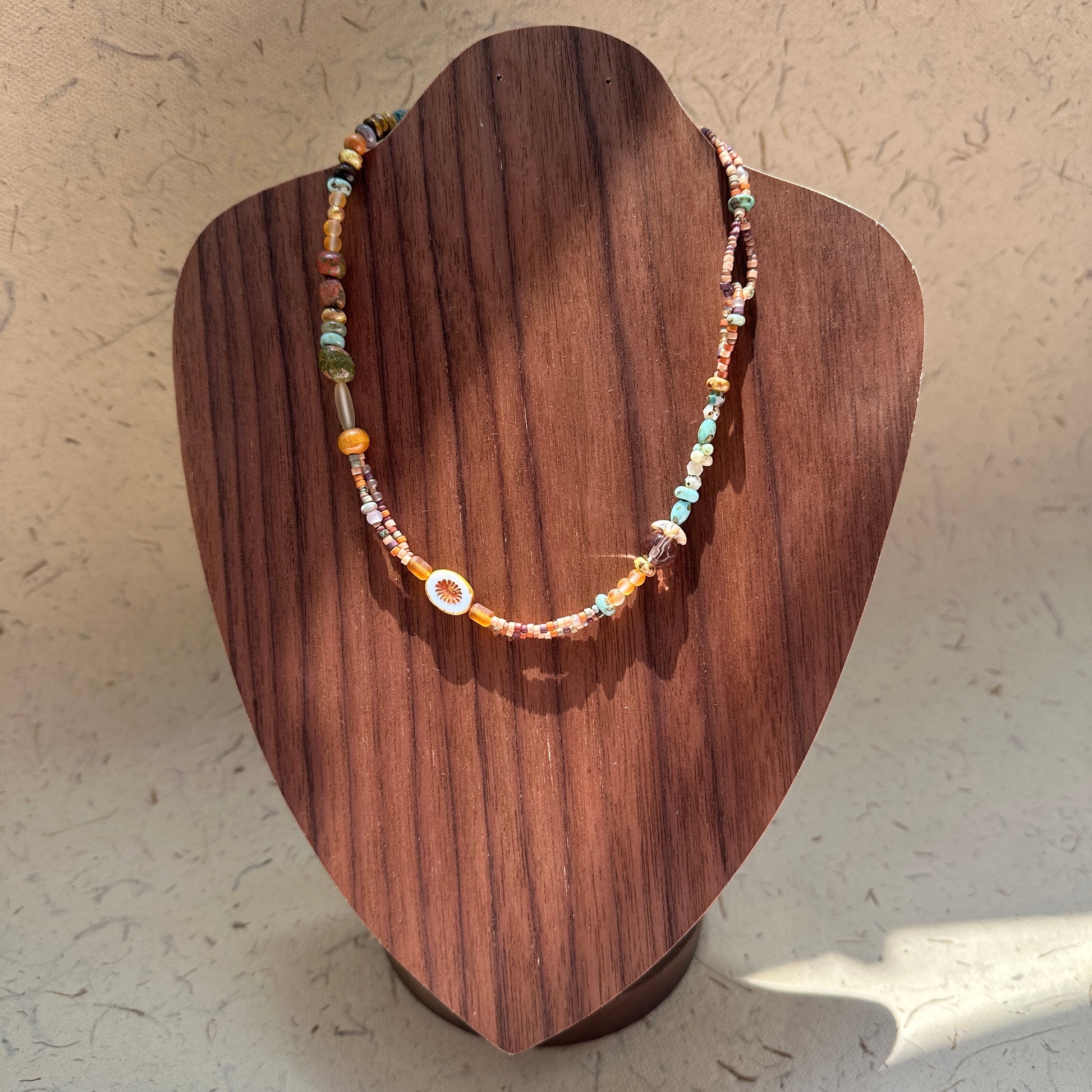 Sunlit Meadow Necklace on wood display bust — warm amber and earthy-green beads with a sunflower-inspired charm