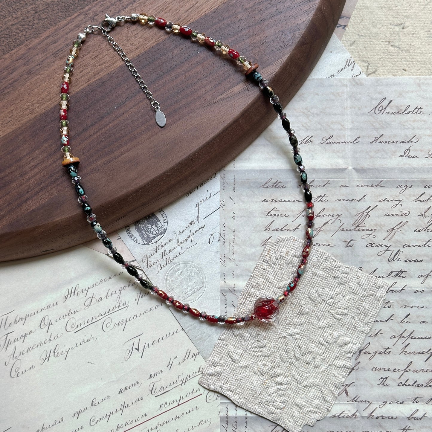 Crimson Eclipse Necklace — flat lay on vintage papers beside a round wooden tray; red, black, and clear glass beads with a crimson orb focal