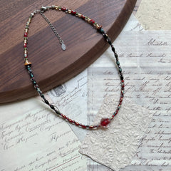 Crimson Eclipse Necklace — flat lay on vintage papers beside a round wooden tray; red, black, and clear glass beads with a crimson orb focal