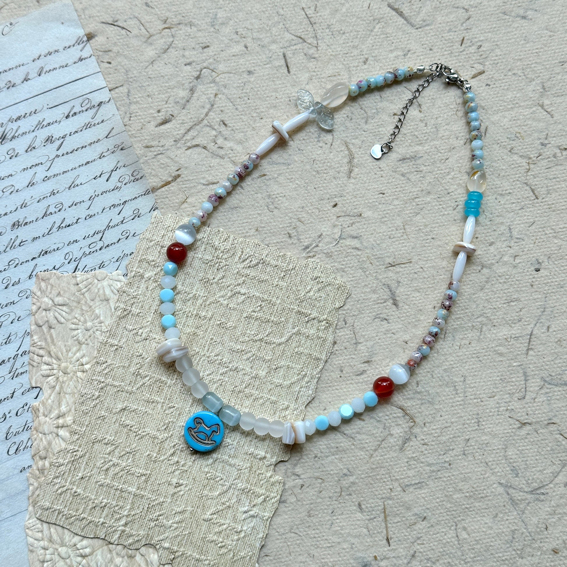 Dolphin Tide Necklace — flat lay on vintage papers; seafoam-blue and pearl-white beads with coral-red accents and a dolphin medallion charm