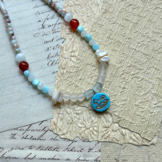 Dolphin Tide Necklace — circle layout on textured paper; coastal palette of seafoam blue, pearl white, and coral red