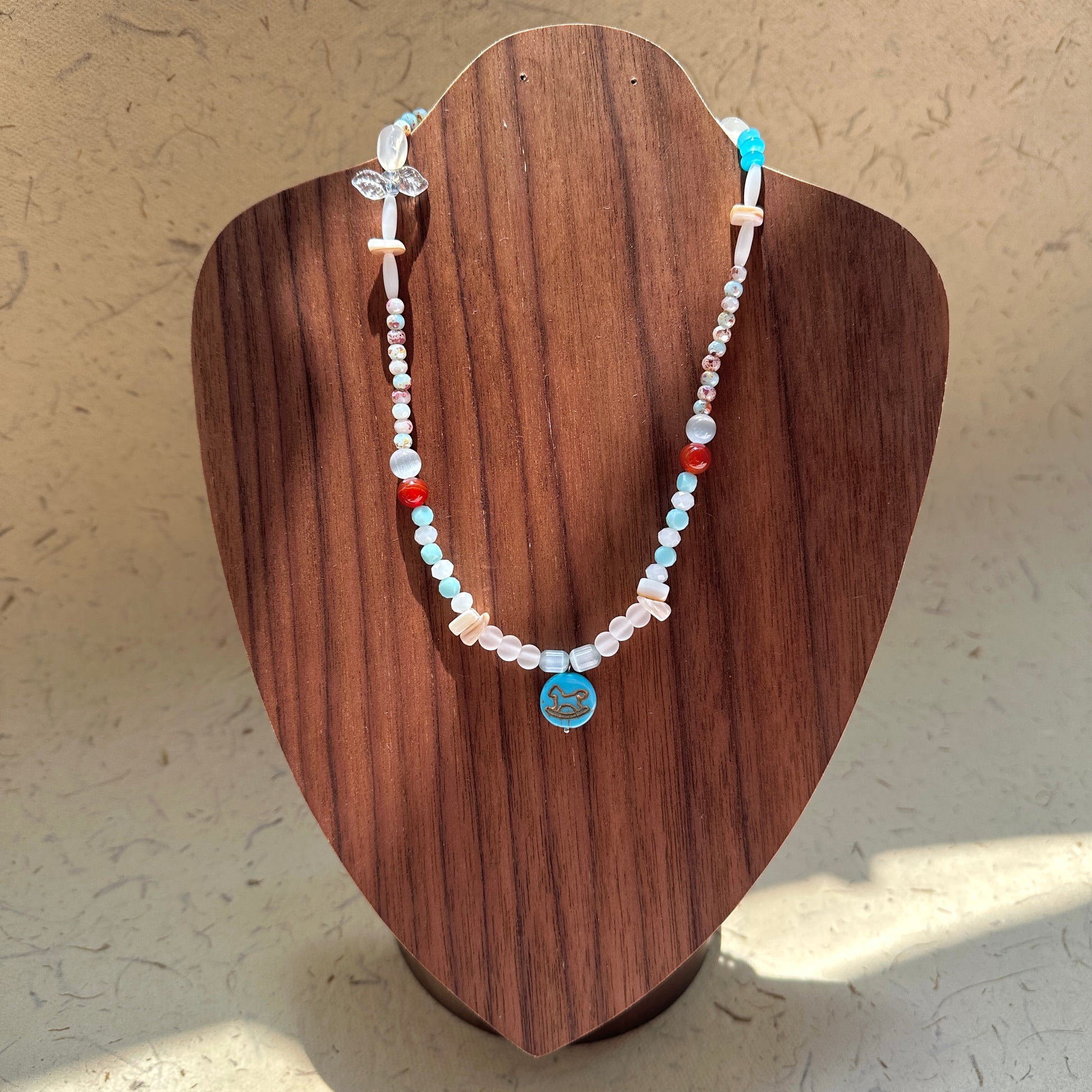 Dolphin Tide Necklace on wood display bust — seafoam-blue and pearl-white beads with a playful dolphin medallion charm