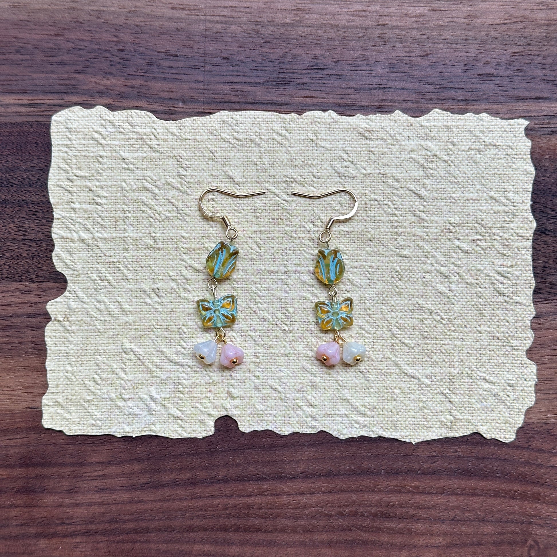 Butterfly Blossom Drops — flat lay on textured cream paper; seafoam tulip beads stacked with butterfly tiles and pastel petal clusters on gold-tone hooks