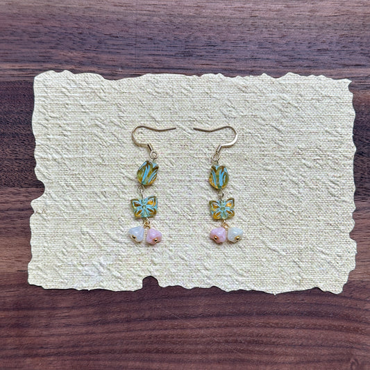 Butterfly Blossom Drops — flat lay on textured cream paper; seafoam tulip beads stacked with butterfly tiles and pastel petal clusters on gold-tone hooks