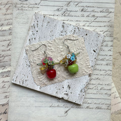 Honeycrisp & Granny Drops — flat lay on textured papers; mismatched red and green glass apples with blossom clusters and silver-tone hooks