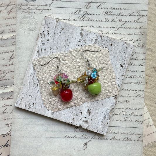 Honeycrisp & Granny Drops — flat lay on textured papers; mismatched red and green glass apples with blossom clusters and silver-tone hooks
