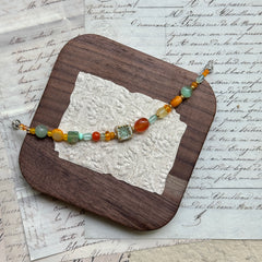 Handmade glass bead bracelet in vibrant citrus hues of orange, yellow, and green with floral charm centerpiece, displayed on a wooden tray with vintage paper background