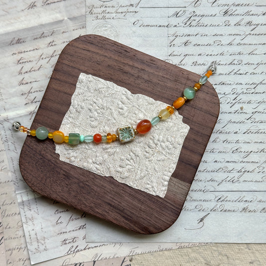 Handmade glass bead bracelet in vibrant citrus hues of orange, yellow, and green with floral charm centerpiece, displayed on a wooden tray with vintage paper background