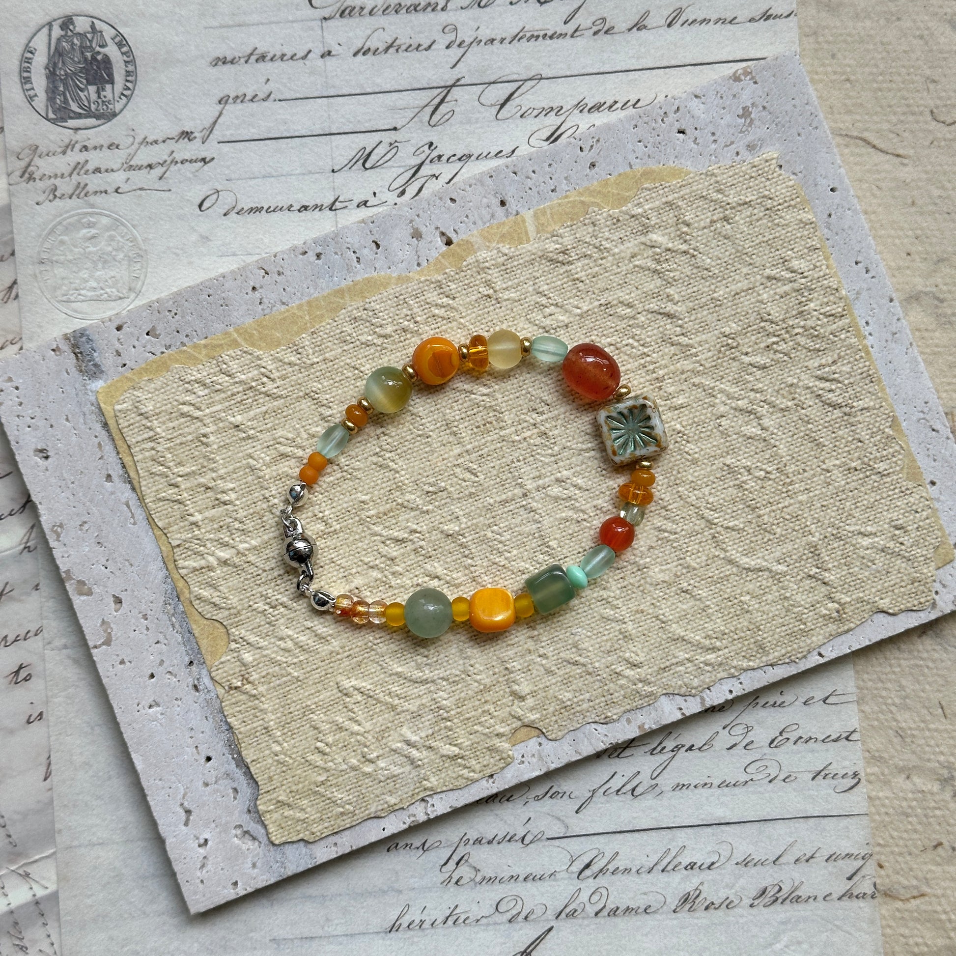 Handmade glass bead bracelet in vibrant citrus hues of orange, yellow, and green with floral charm centerpiece, displayed on a wooden tray with vintage paper background 2