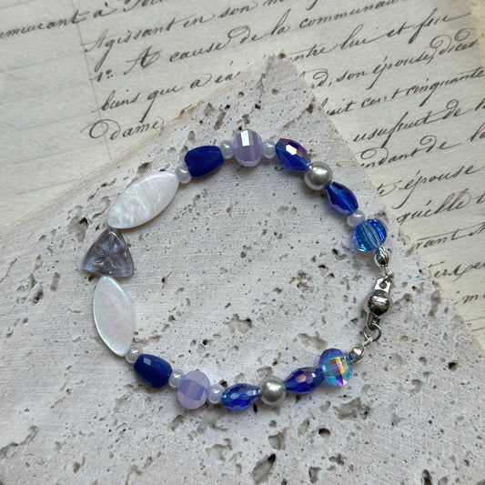 Handcrafted Midnight Pearl Bracelet with deep sapphire blue and pearl-white glass beads, accented by lilac details and floral motif centerpiece, displayed on textured paper background.