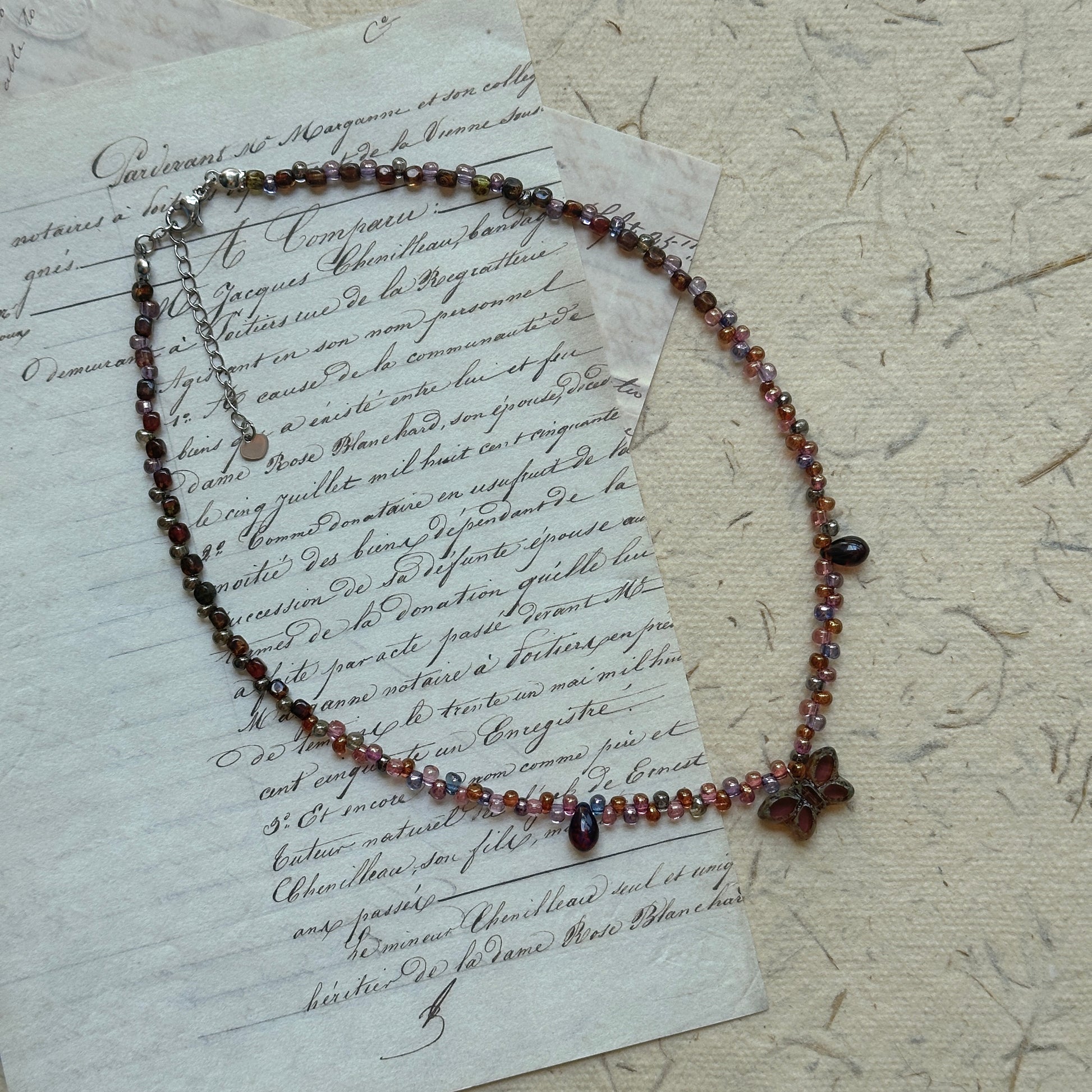 Twilight Bloom Necklace by Glazenya – handcrafted beaded necklace in wine-red, plum, and amber tones with a floral charm, displayed on vintage letter background.