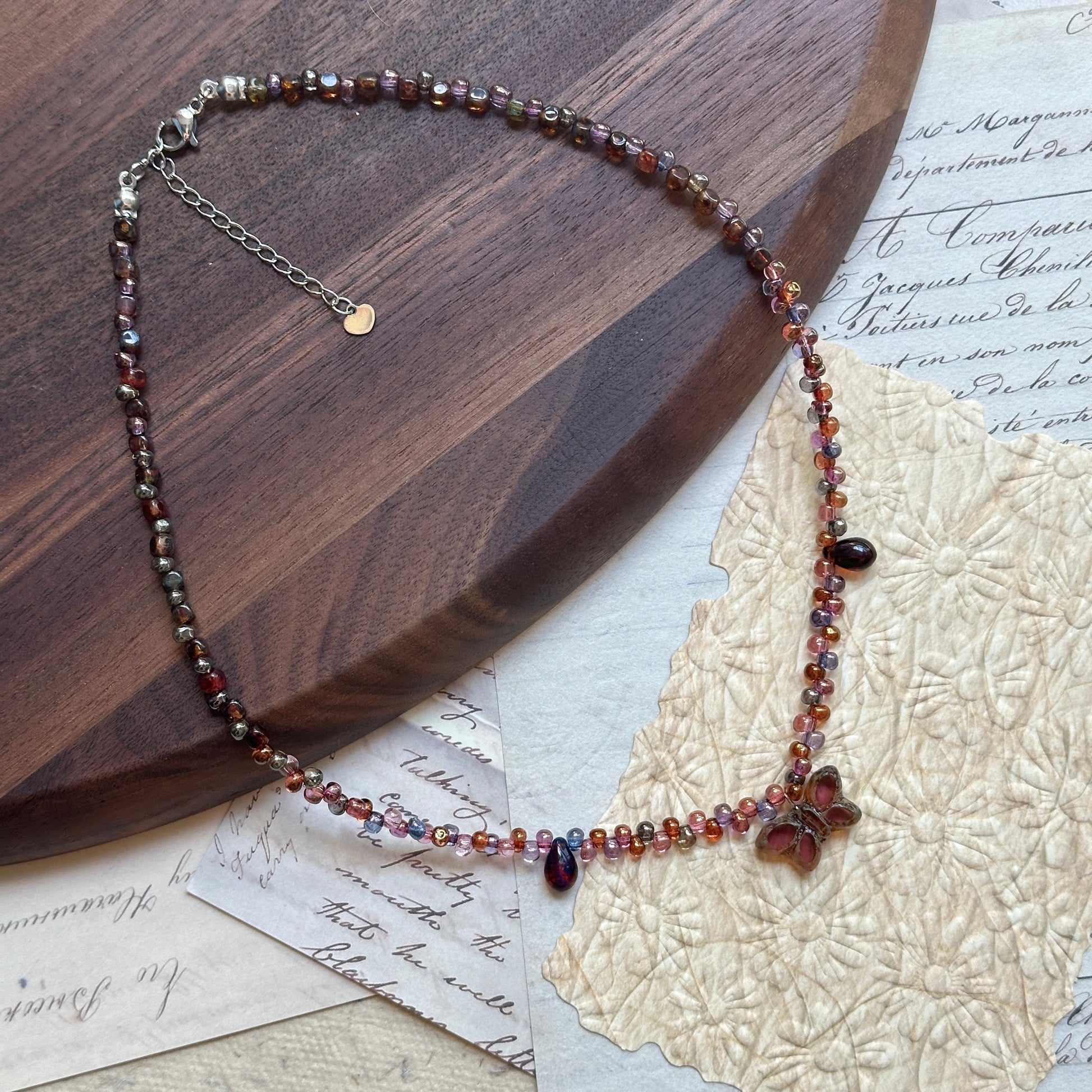 Close-up of Glazenya Twilight Bloom Necklace – plum and amber glass beads with delicate flower charm on wood display.