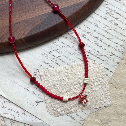 Crimson Toadstool Necklace on wooden surface, featuring red beads with ivory accents and a whimsical lampwork mushroom pendant