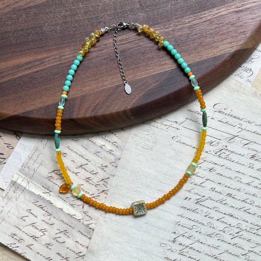 Citrus Lagoon Necklace displayed on wooden surface, featuring floral ceramic centerpiece, citrus yellow and lagoon green beads, and amber leaf charm