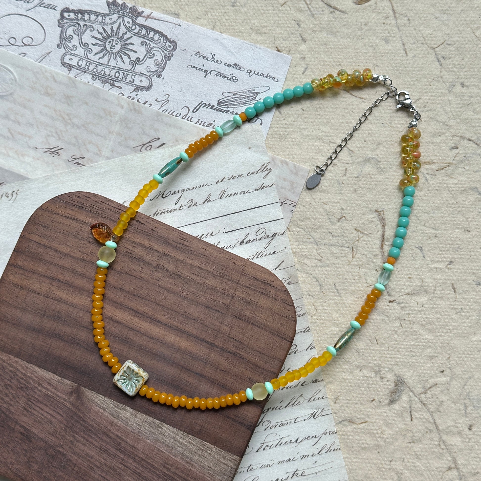 Full view of Citrus Lagoon Necklace laid flat, highlighting colorful bead palette, ceramic flower centerpiece, and amber leaf accent