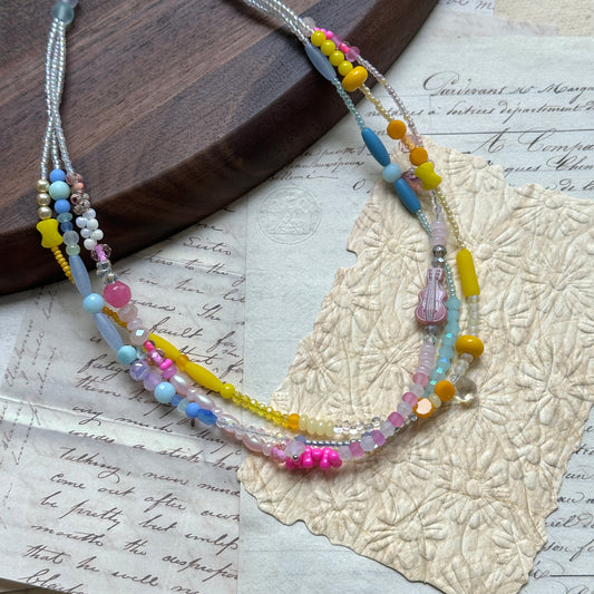 Close-up of Sunshine Confetti Necklace on wooden surface, featuring layered pastel beads in yellow, blue, pink, and purple with crystal accents