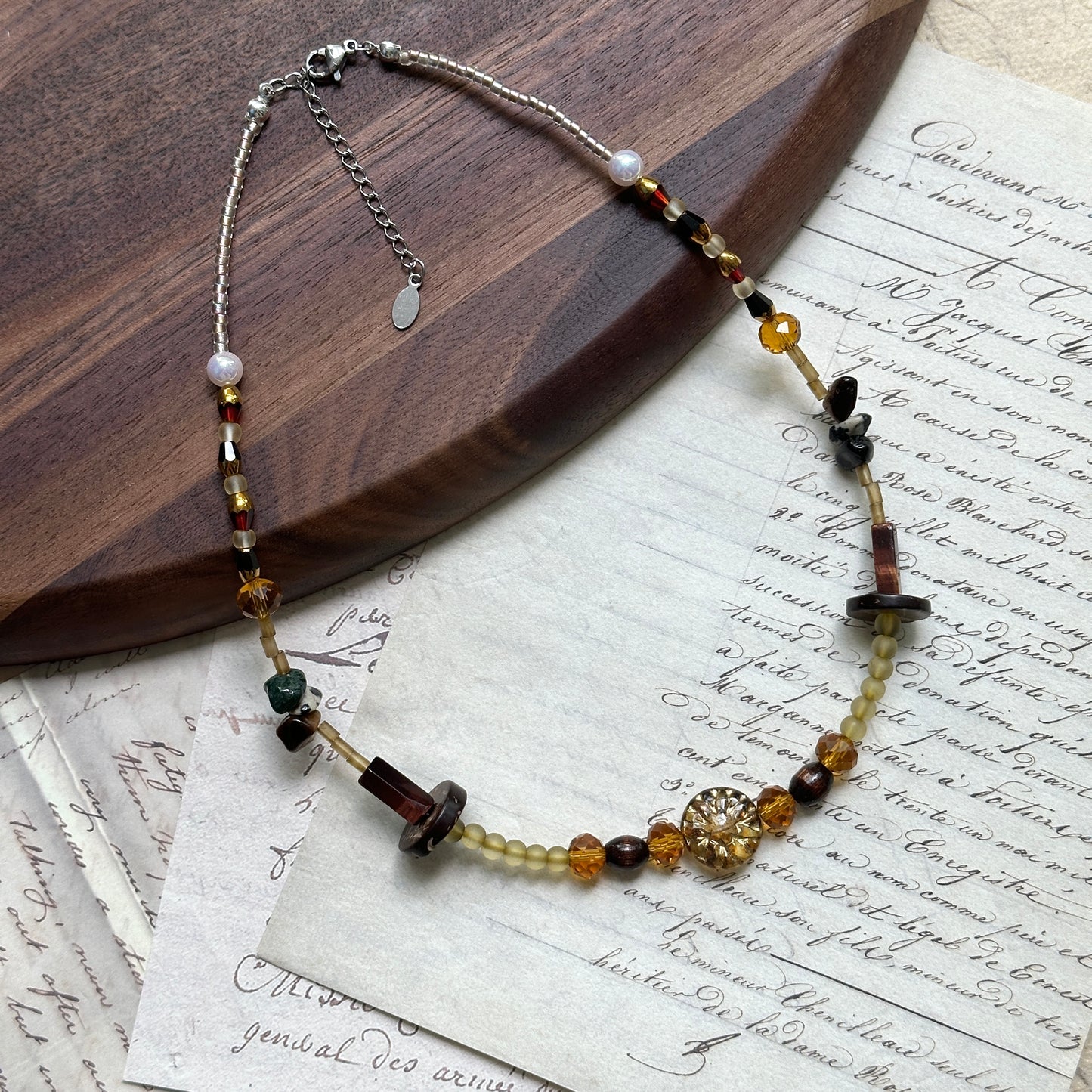 Marigold Ember Necklace close-up on wooden surface, featuring golden sunburst centerpiece, amber-yellow beads, moss-green stones, and deep brown accents