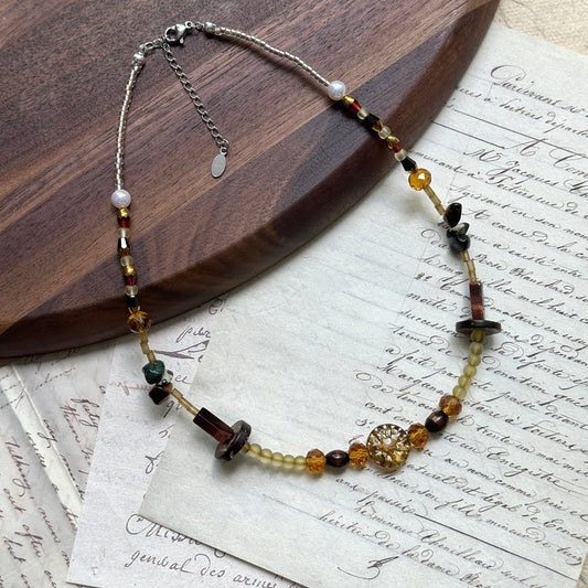 Marigold Ember Necklace close-up on wooden surface, featuring golden sunburst centerpiece, amber-yellow beads, moss-green stones, and deep brown accents