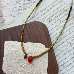 Wild Strawberry Necklace close-up on rustic paper, featuring glass strawberry pendant with petite green leaf, dewdrop accent, and moss-green beads
