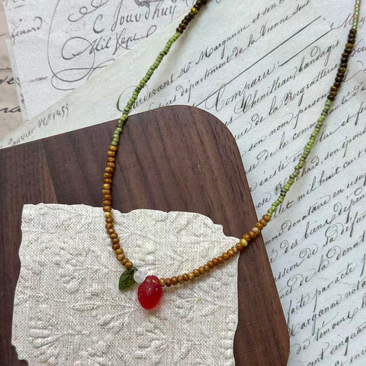 Wild Strawberry Necklace close-up on rustic paper, featuring glass strawberry pendant with petite green leaf, dewdrop accent, and moss-green beads
