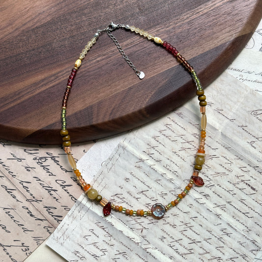 Olive Grove Blossom Necklace close-up on wooden surface, featuring floral glass coin pendant, olive-green beads, honey-amber accents, and brown tones