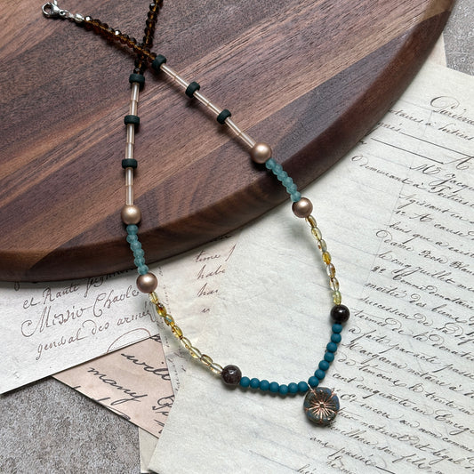 Lagoon Sunburst Necklace close-up on rustic surface, featuring lagoon-blue seed beads, champagne pearls, and radiant sunburst pendant