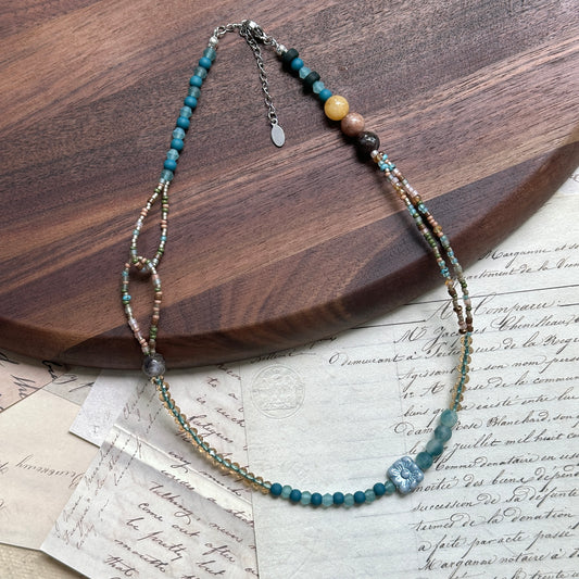Aqua Loop Blossom Necklace close-up on wooden surface, featuring lagoon-blue beads, sandy-gold shimmer, and floral glass focal