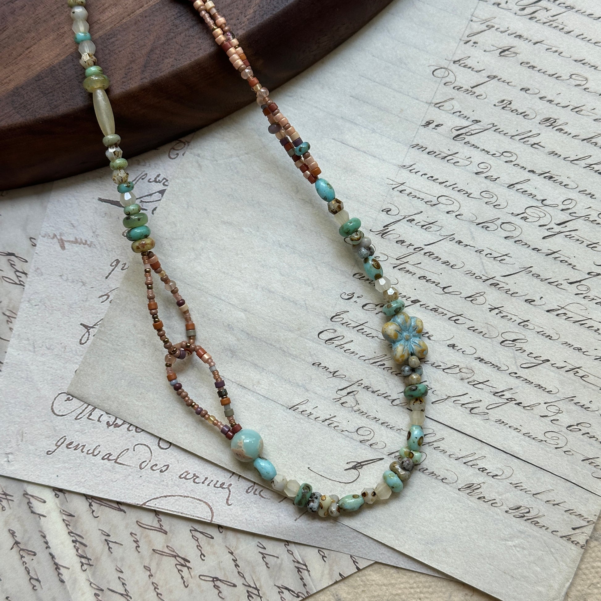 Sage Loop Blossom Necklace close-up on rustic paper, featuring floral Czech-glass focal, sage and mint beads, sandy peach tones, and double-loop detail