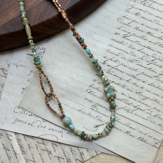 Sage Loop Blossom Necklace close-up on rustic paper, featuring floral Czech-glass focal, sage and mint beads, sandy peach tones, and double-loop detail