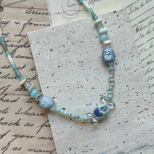Ocean Whisper Necklace close-up on textured paper, featuring aqua blue and pearl-white beads with whimsical glass charms of penguin and jellyfish