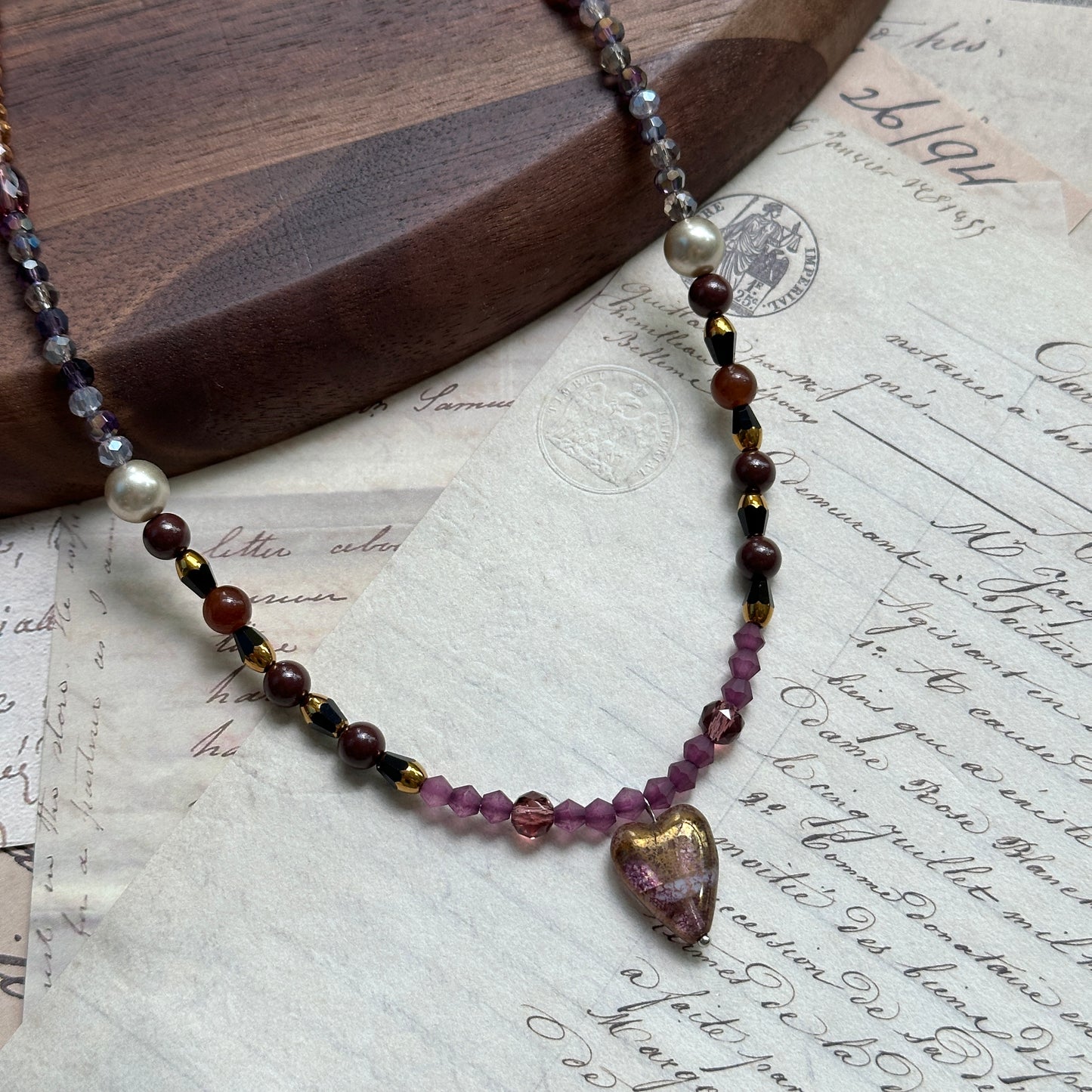 Amour Heart Necklace close-up on rustic surface, featuring blush-pink glass heart pendant with gradient burgundy, plum, and gold beads