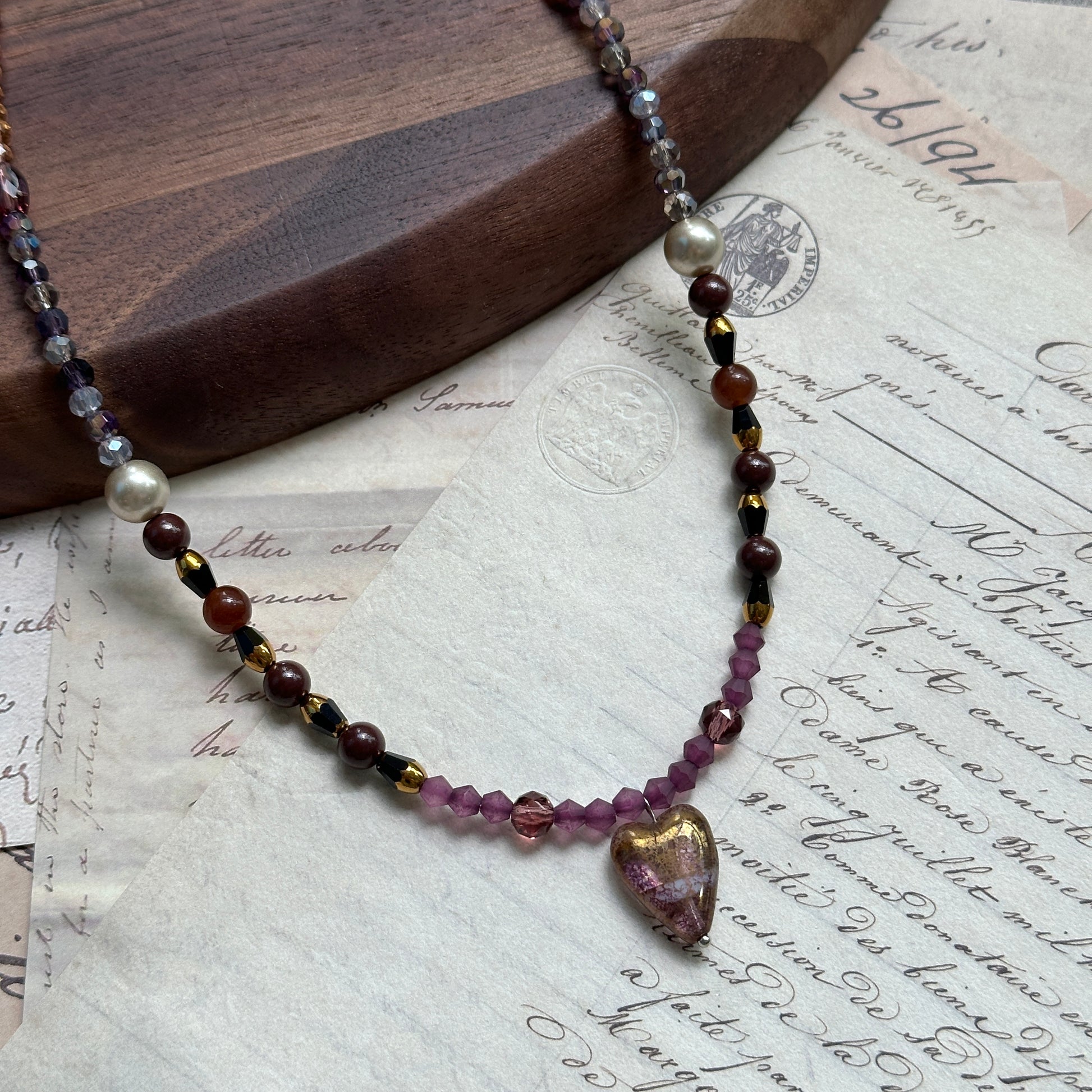 Amour Heart Necklace close-up on rustic surface, featuring blush-pink glass heart pendant with gradient burgundy, plum, and gold beads