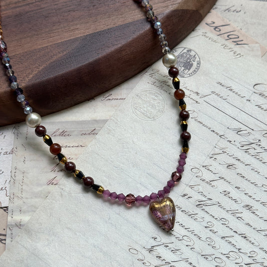 Amour Heart Necklace close-up on rustic surface, featuring blush-pink glass heart pendant with gradient burgundy, plum, and gold beads