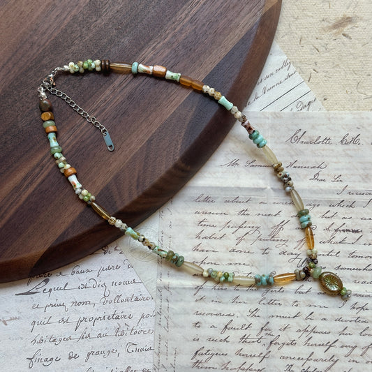 Verdant Amulet Necklace close-up on wooden surface, featuring amulet-style pendant with sage green, amber-gold, and ivory beads