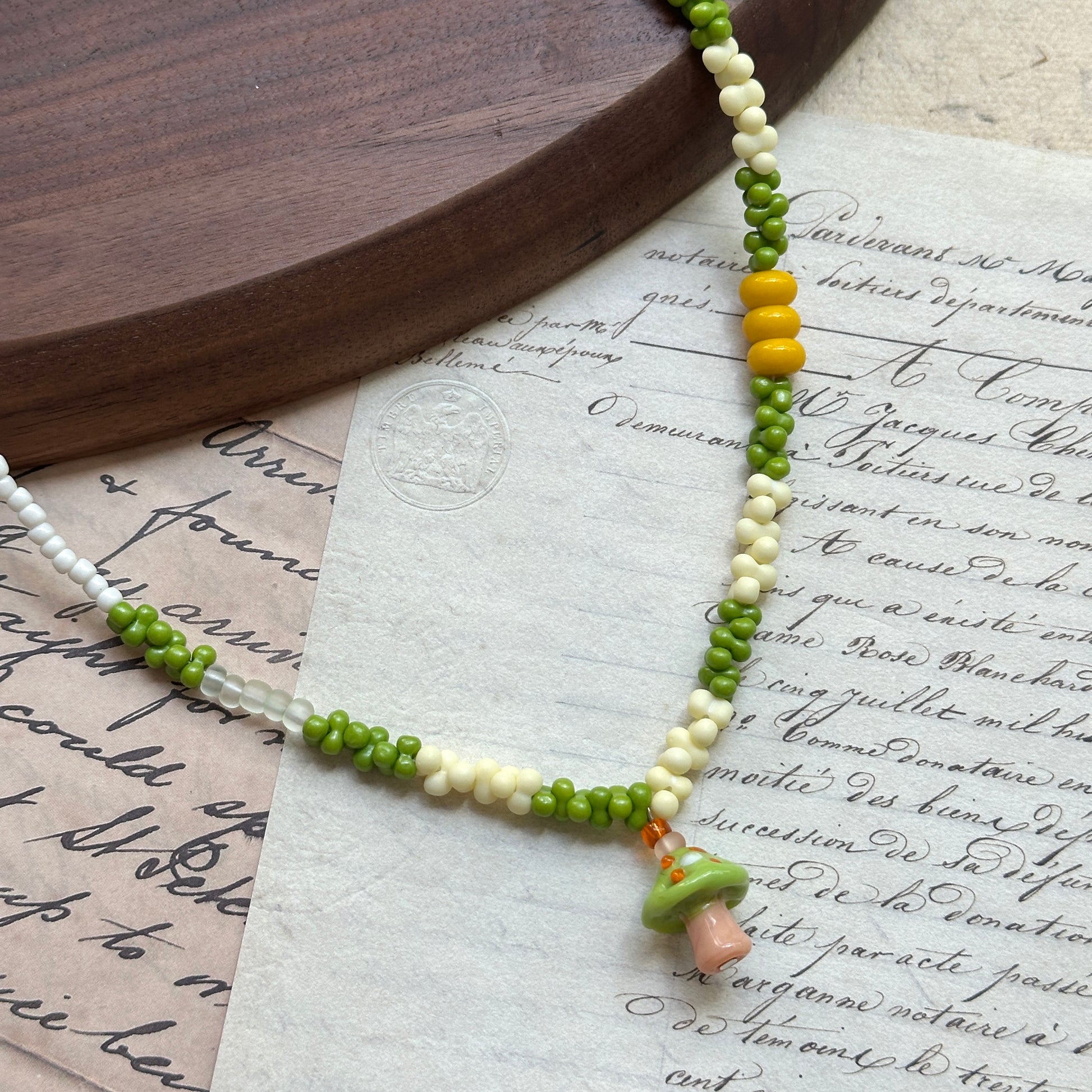 Forest Mushroom Necklace close-up on rustic paper, featuring whimsical glass mushroom pendant in green and peach with yellow and ivory beads