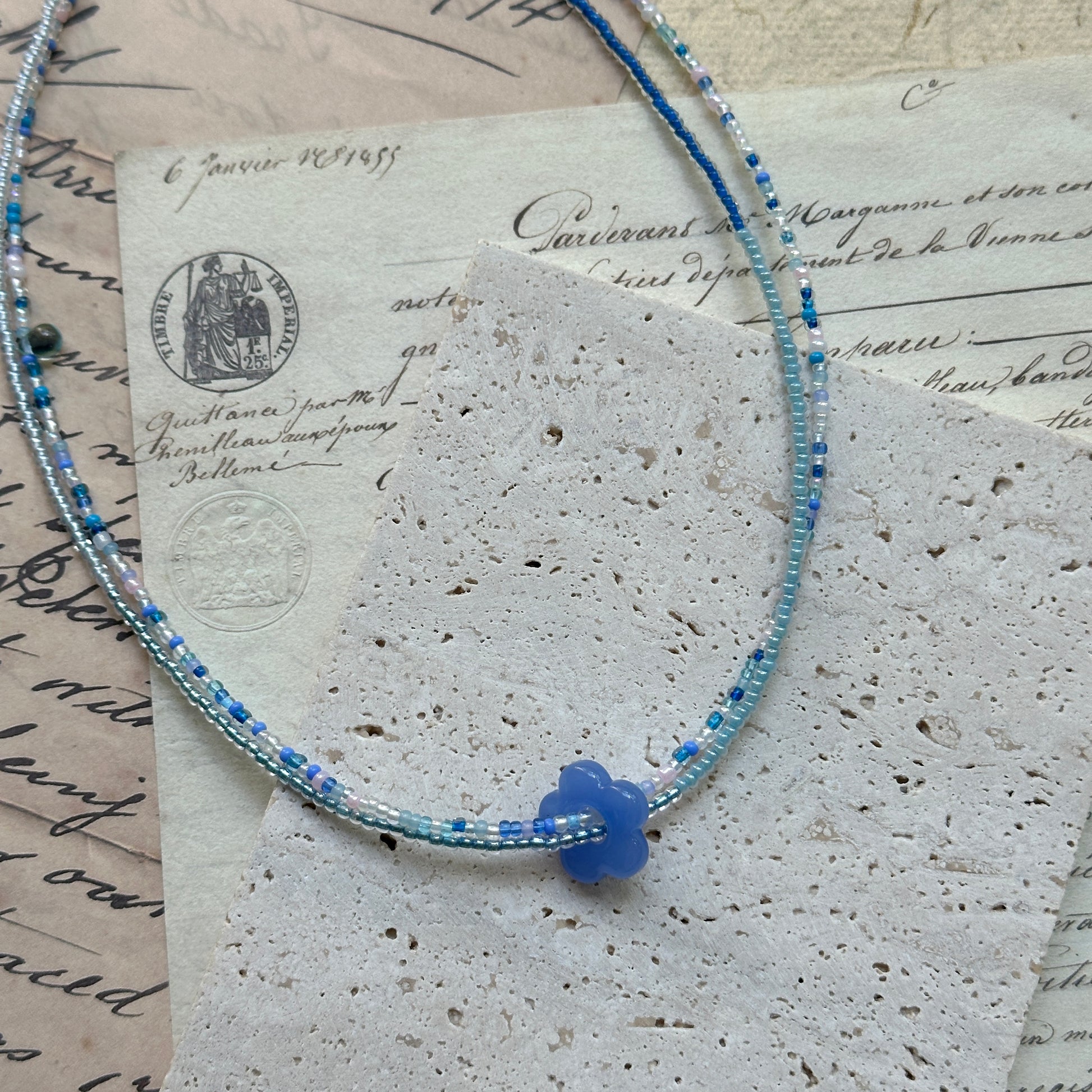 Azure Bloom Necklace close-up on rustic paper, featuring petite azure glass flower pendant with delicate strands of sky blue and pearl white beads